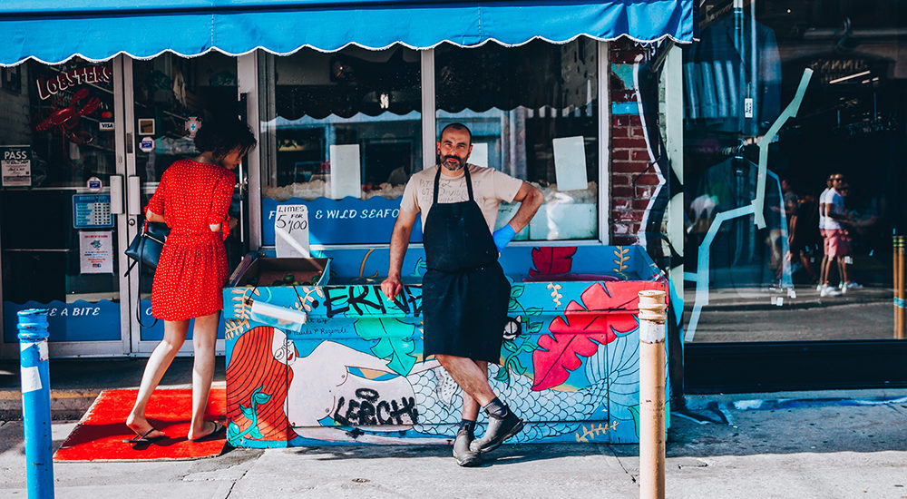 Herman standing outside of Hooked whilst a woman is shopping for fish.