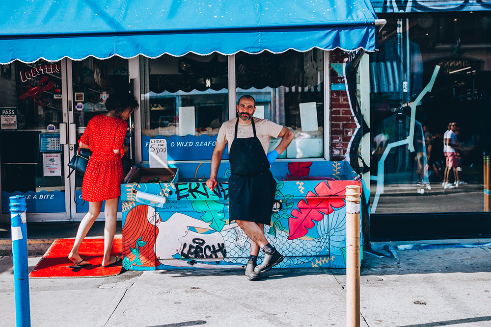 Herman standing outside of Hooked whilst a woman is shopping for fish. 